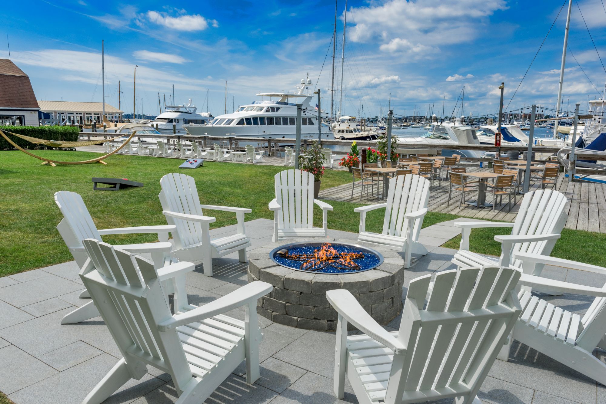 Outdoor seating with white chairs around a fire pit by a marina, featuring boats and a hammock under a blue sky.