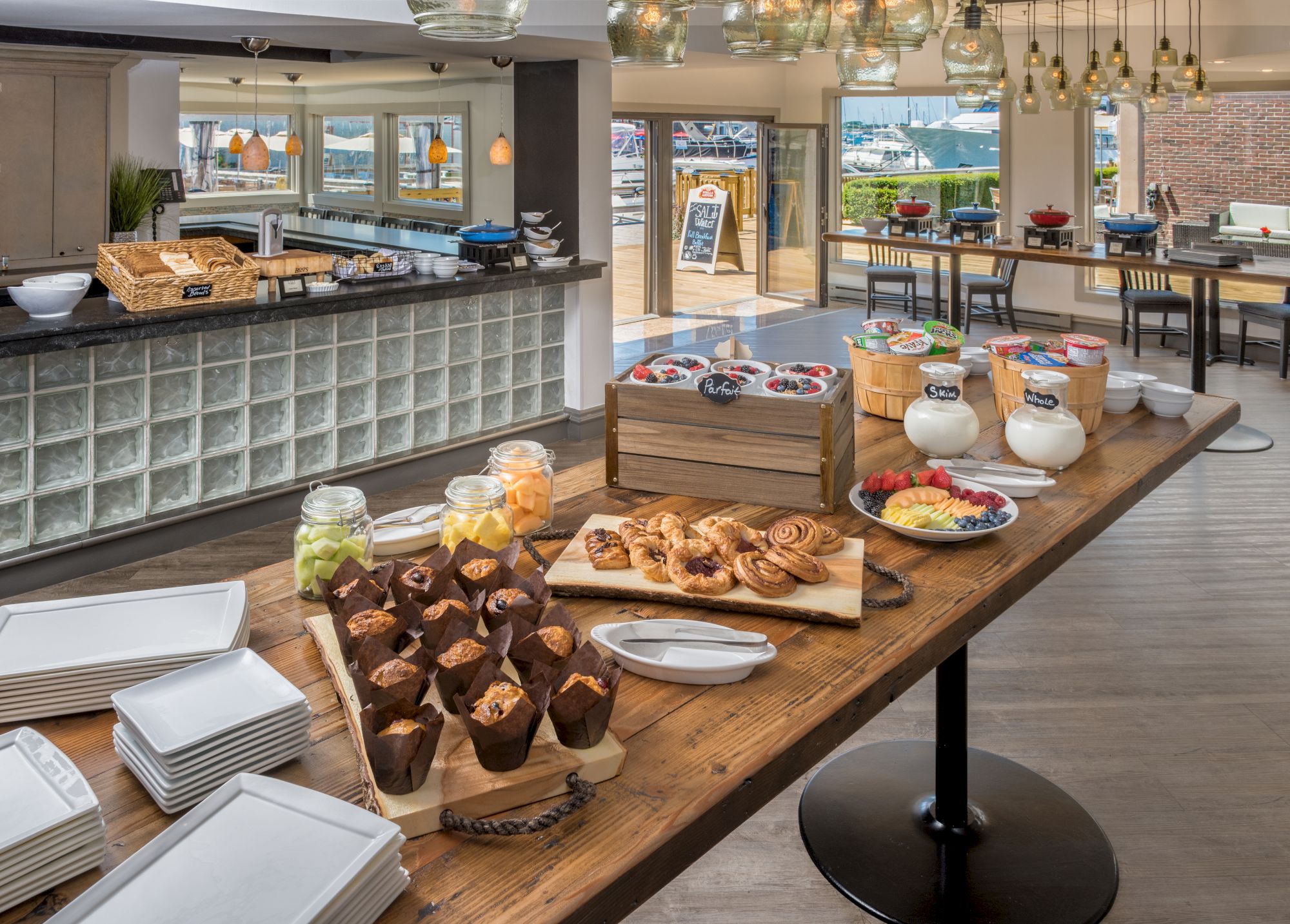 A breakfast buffet with pastries, muffins, fruits, and cereals is set up in a modern dining area with large windows and decorative lights.