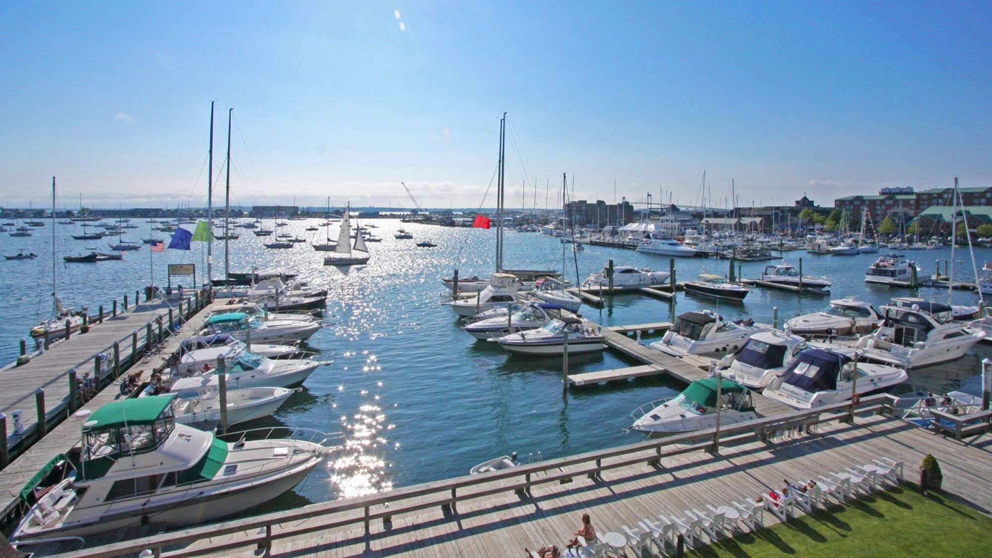 A marina with numerous boats docked on a sunny day, surrounded by calm water and a clear blue sky.