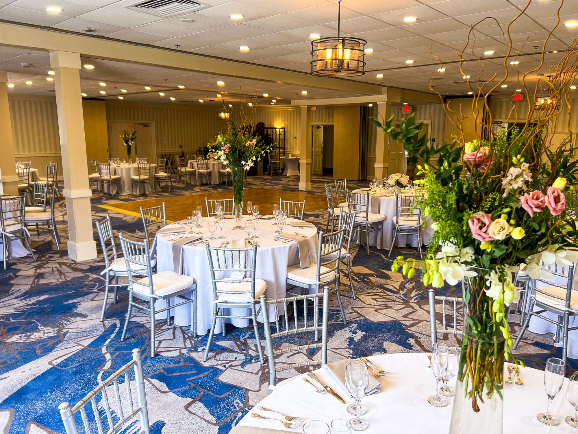 An elegantly set banquet hall with round tables, white tablecloths, floral centerpieces, and silver chairs arranged for an event.