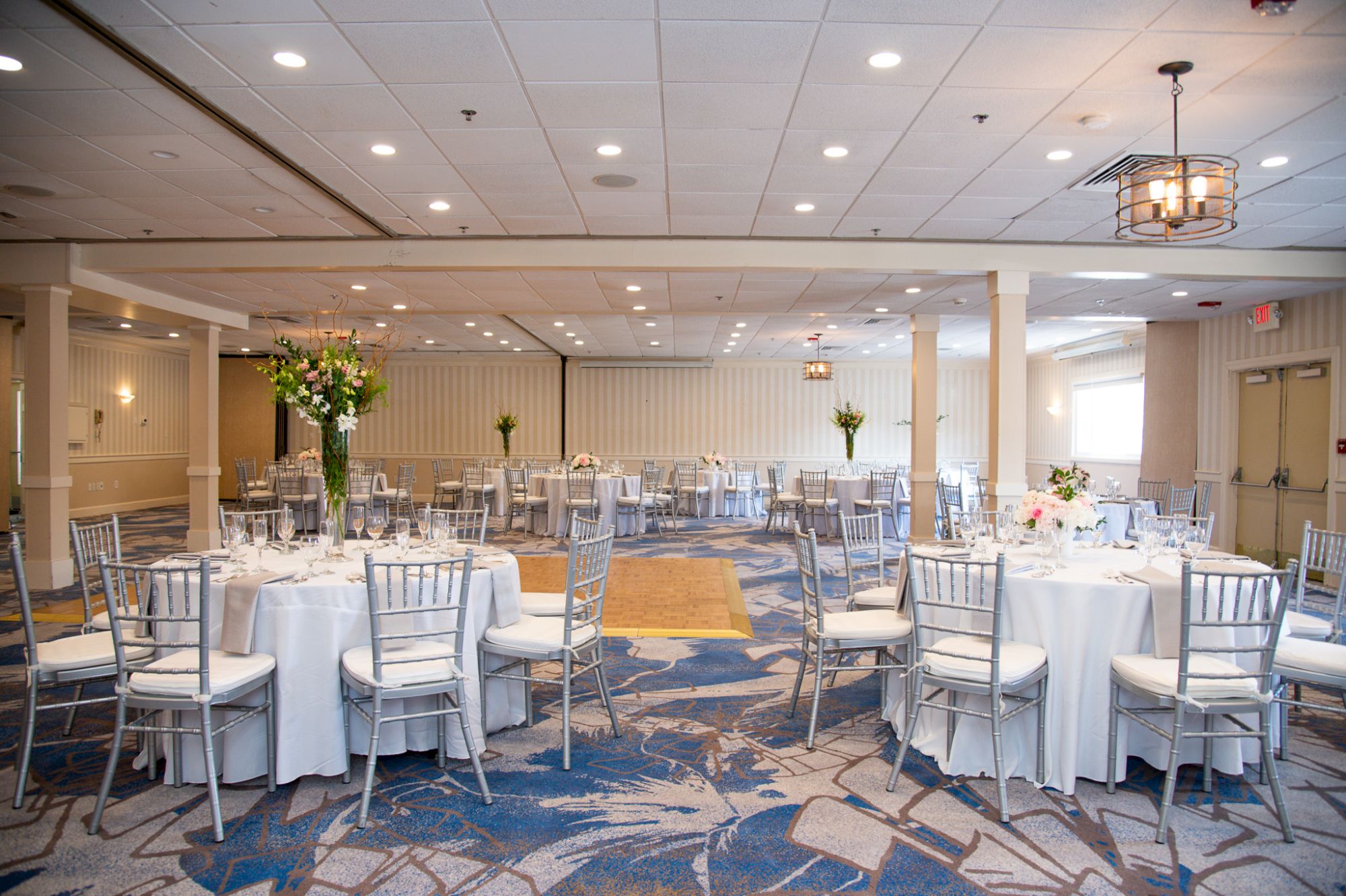 A decorated banquet hall with round tables, white linens, silver chairs, and floral centerpieces, ready for an event.