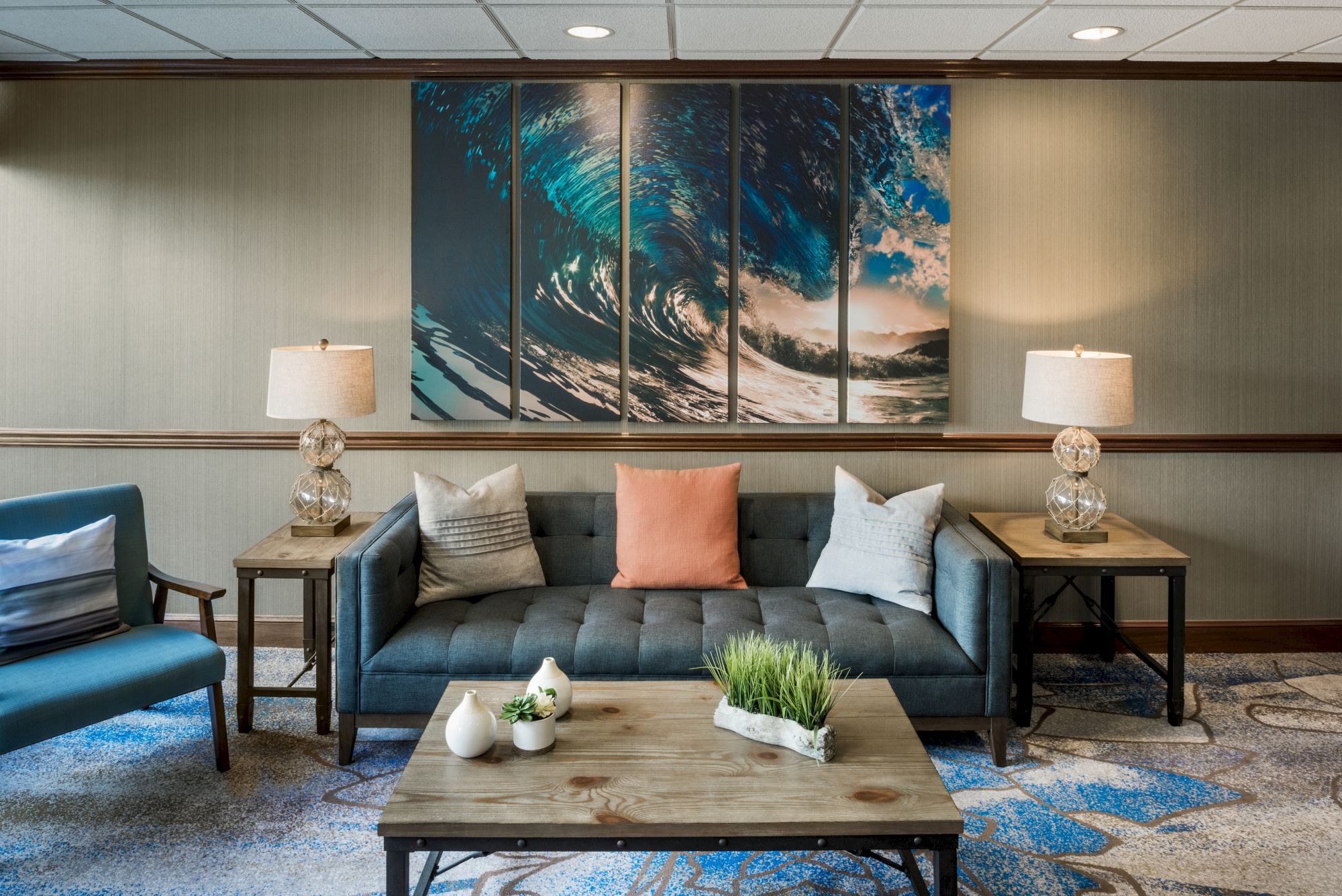 A stylish lounge with a blue sofa, colorful pillows, wooden tables, lamps, and ocean-themed artwork on the wall.