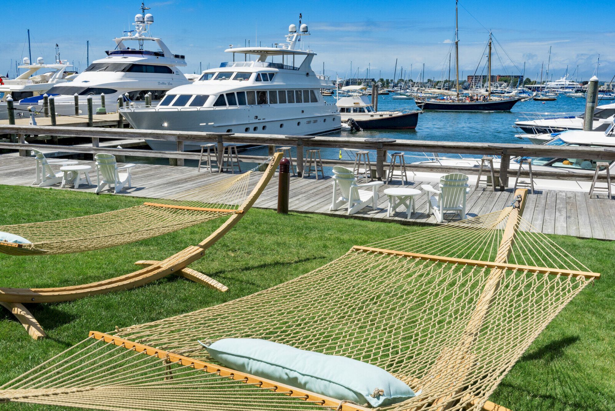 The image shows a marina with yachts docked, a grassy area with hammocks, and a wooden deck with chairs and tables under a clear blue sky.