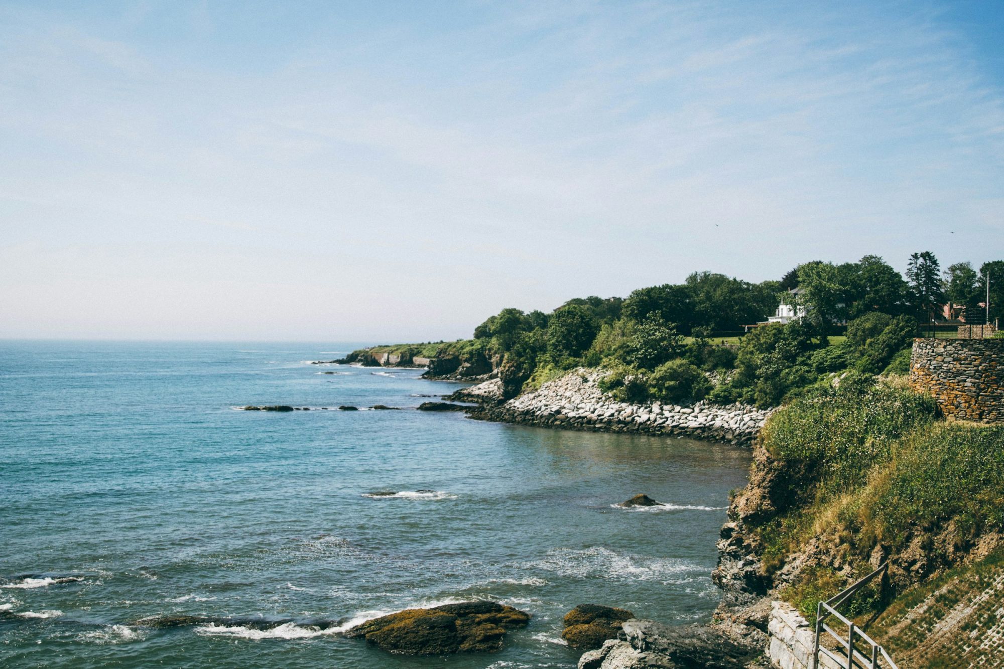 A coastal landscape featuring rocky cliffs, lush greenery, and calm blue ocean waters under a clear sky.