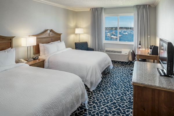 A hotel room with two beds, a TV, a desk, and a window showing a view of boats on the water.