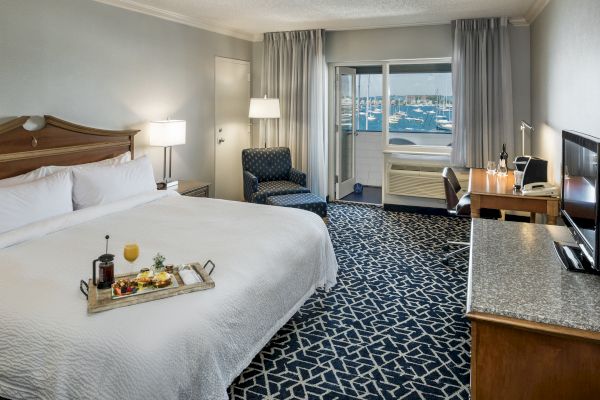 A hotel room with a large bed, desk, TV, armchair, and a tray of breakfast on the bed. Balcony shows a marina view.