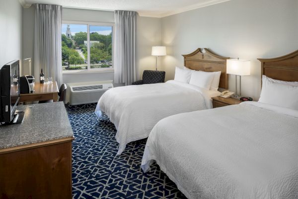 The image shows a hotel room with two beds, a window view, a TV, desk, and lamps on nightstands, creating a cozy and clean atmosphere.