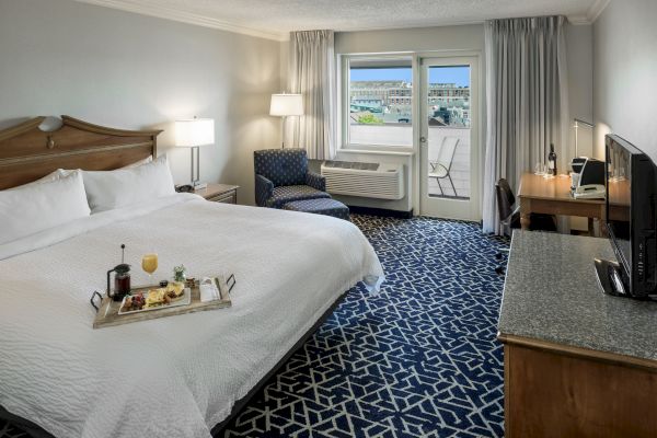 A hotel room with a large bed, TV, desk, armchair, and a tray with drinks. There's a window with a view. Navy patterned carpet adds style.