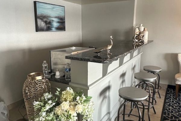 The image shows a home bar area with stools, decorative vases, and flowers, set against a gray wall with a picture hanging above.