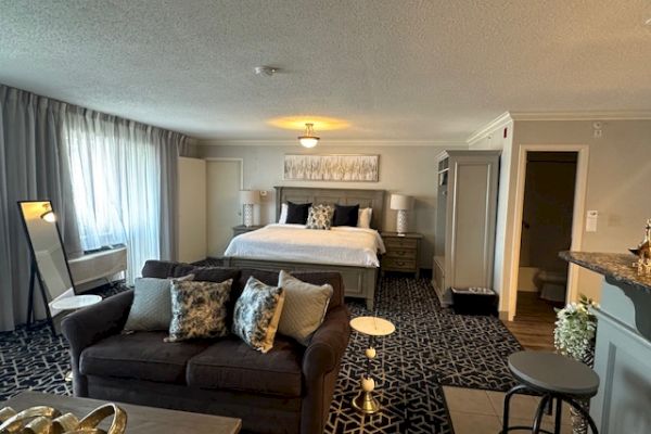 A modern hotel room with a bed, sofa, chair, and bar stools. There’s a patterned carpet and soft lighting.