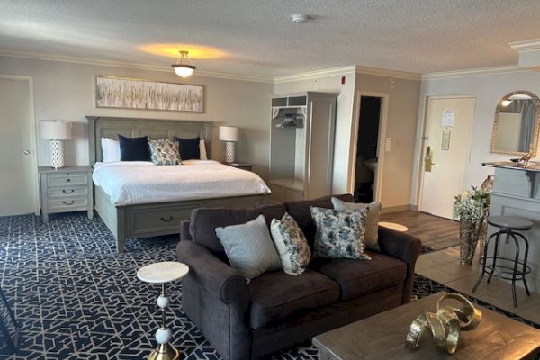 A spacious hotel room with a bed, sofa, and coffee table. It features modern decor, a patterned rug, and neutral color tones.