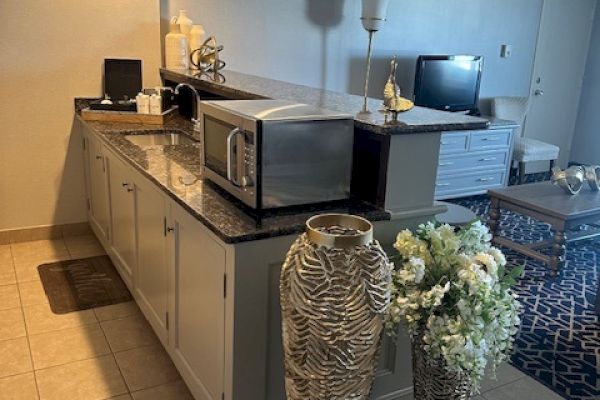 A kitchen area with a countertop, microwave, large decorative vases, flowers, and a living room with a TV in the background.