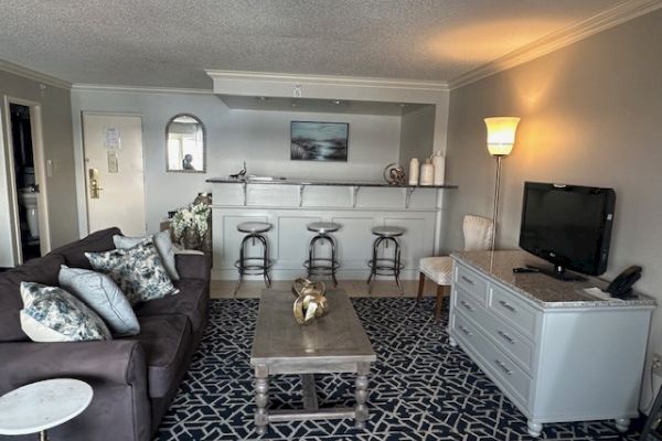 A cozy living room with a sofa, coffee table, TV, bar stools, and a kitchenette. Decorative pillows and a patterned rug add style.