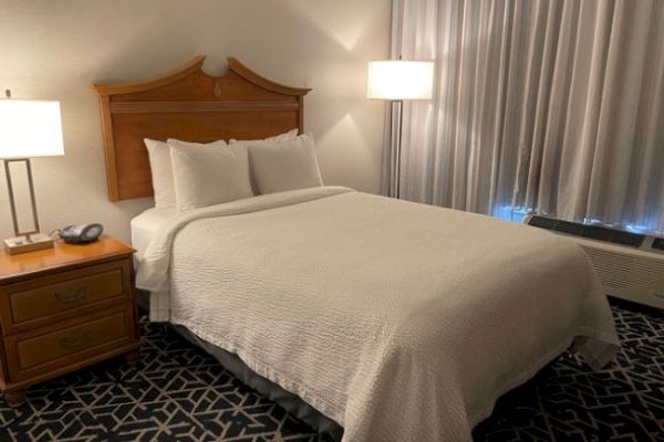 A neatly made bed with white linens is in a hotel room, featuring wooden furniture, lamps, and patterned carpet flooring.