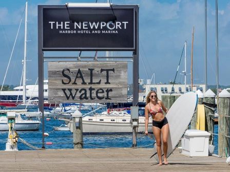 A woman in a bikini holds a surfboard on a dock near boats. Sign reads "THE NEWPORT Harbor Hotel and Marina" and "SALT water."