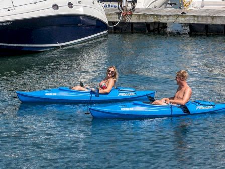 Two people are kayaking in blue kayaks on calm water near white boats at a marina, enjoying a sunny day.