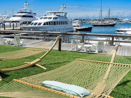 A marina with docked yachts, loungers, and hammocks on a grassy area overlooking the water. Sunny skies and a relaxing atmosphere.