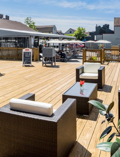 The image shows a sunny outdoor patio with wicker furniture, glass tables, and potted plants, surrounded by brick buildings and a wooden deck.