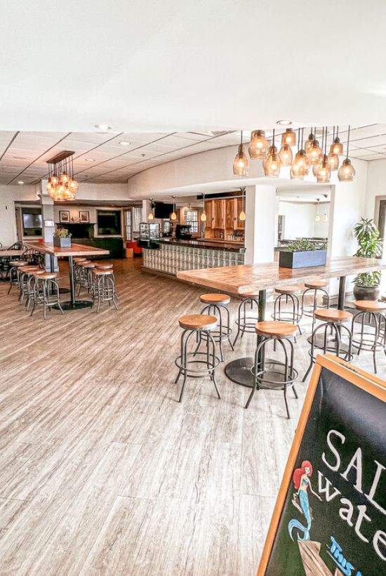 The image shows a bright, modern cafe interior with wooden floors, tables, chairs, hanging lights, and a chalkboard sign reading 