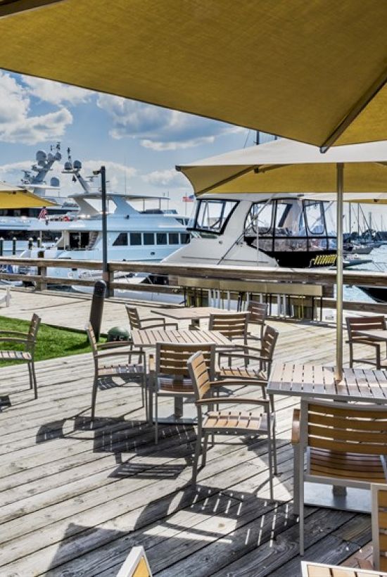 A marina with boats docked, outdoor seating under umbrellas on a wooden deck, and a grassy area under a partly cloudy sky.