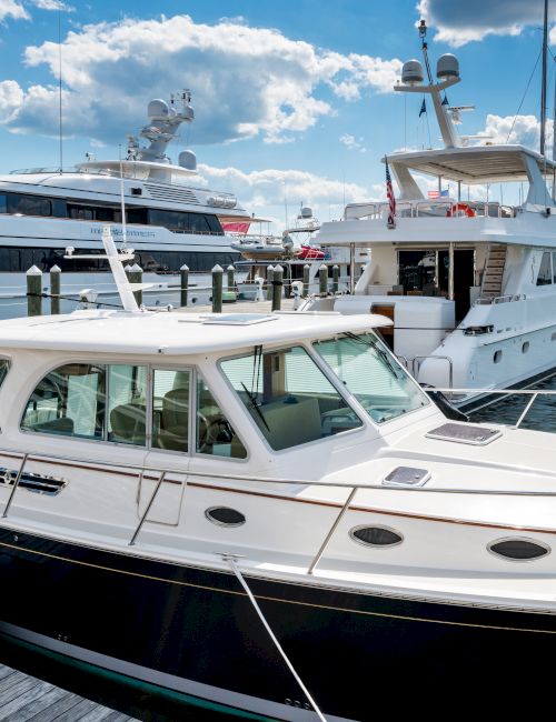 A marina scene with several yachts docked alongside a wooden pier, under a blue sky with scattered clouds.