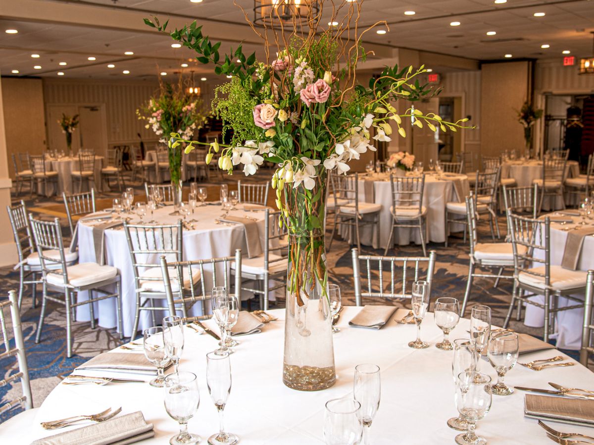 The image shows an elegant banquet hall setup with round tables, white tablecloths, floral centerpieces, and neatly arranged chairs.