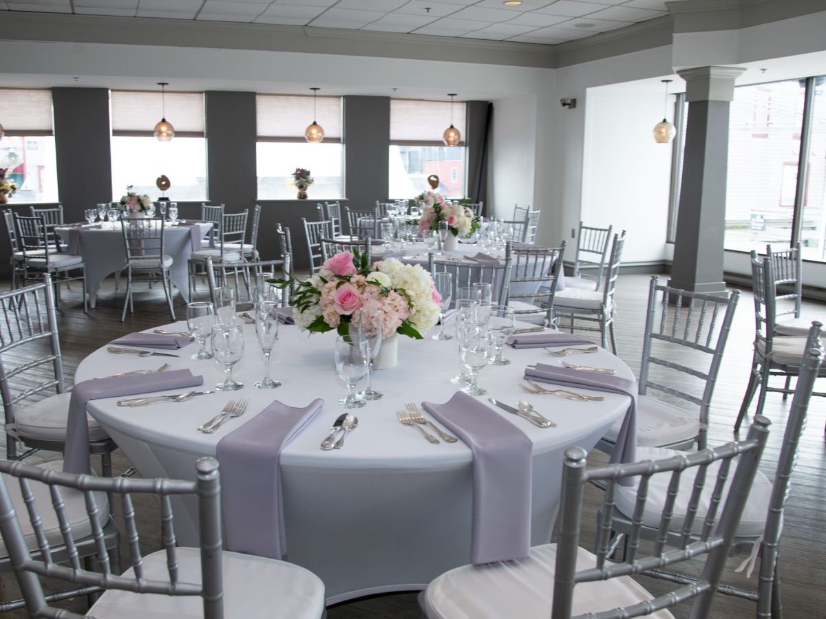 An elegant banquet hall setup with round tables, white tablecloths, pastel napkins, and floral centerpieces, surrounded by silver chairs.