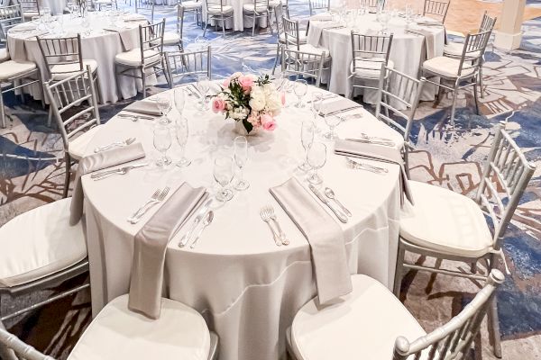 The image shows an elegant banquet hall set up with round tables, white tablecloths, silver chairs, and flower centerpieces.