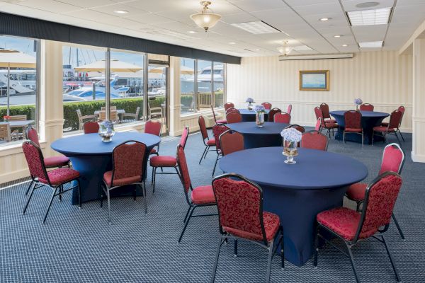 A conference room with round tables covered in blue tablecloths and red chairs, overlooking a view of boats outside large windows.