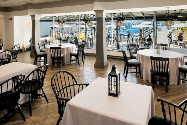 A restaurant with white tablecloths and black chairs, featuring large windows overlooking a waterfront.