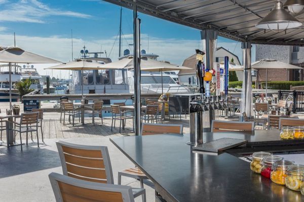 An outdoor restaurant bar with wooden chairs overlooks a marina with docked boats. The area is shaded by umbrellas and a metal awning.