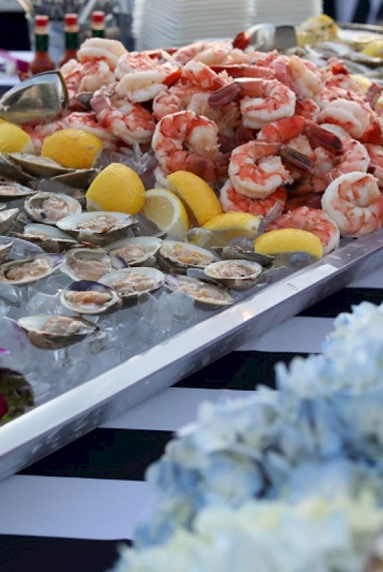 The image shows a seafood platter with shrimp, clams, and lemon wedges, decorated with flowers and pineapple, on a striped tablecloth.