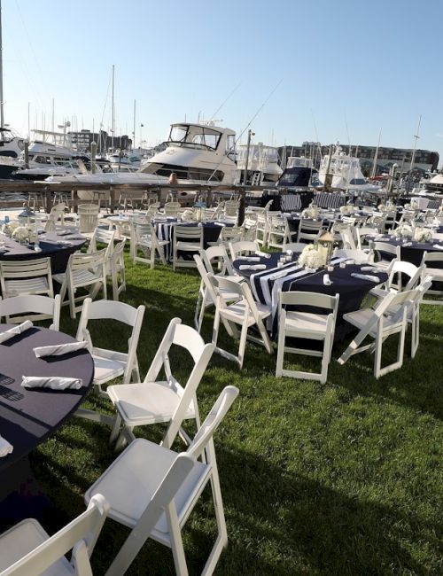 Outdoor event setup with white chairs and tables covered in navy tablecloths by a marina with boats under a clear sky.