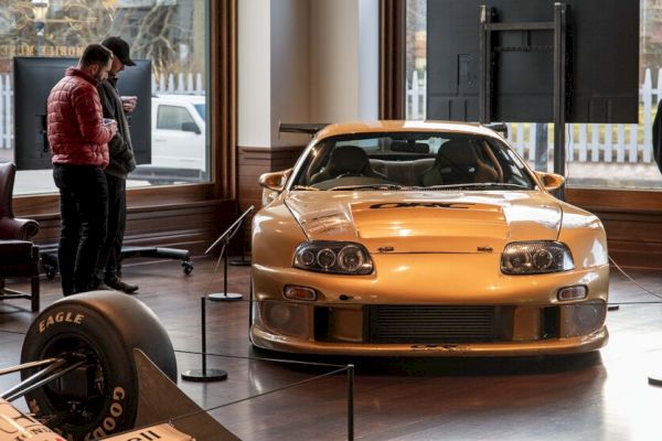 A gold sports car is on display in a showroom with two people observing it. Another vehicle's front section appears on the left.