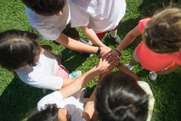 A group of children standing in a circle, each placing their hand in the center on top of the others, on a grassy field.