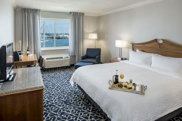 A hotel room features a king-sized bed, a tray with breakfast, a TV on a dresser, a window with a marina view, and a patterned carpet.