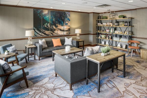 A modern lounge with gray sofas, wooden tables, a bookshelf, and ocean-themed artwork on the wall, accented by decorative plants.