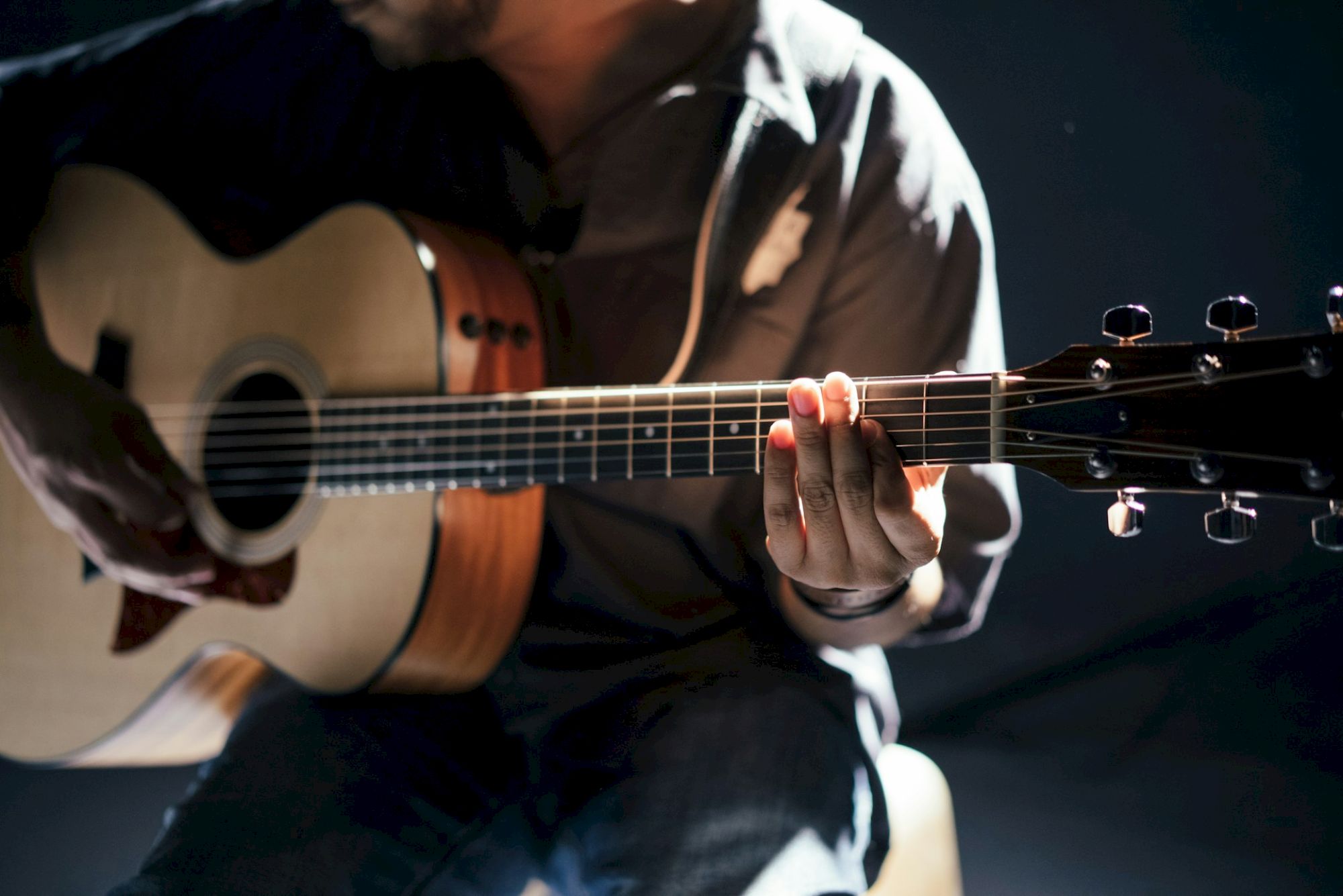 The image shows a person playing an acoustic guitar, with focus on their hand on the fretboard, under dramatic lighting.