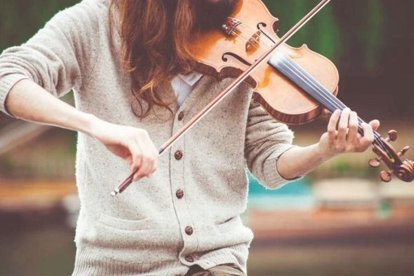 A person playing a violin outdoors, wearing a sweater, and focusing on the instrument.