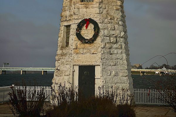 A stone lighthouse is decorated with a holiday wreath, standing near the water with a cloudy sky in the background.