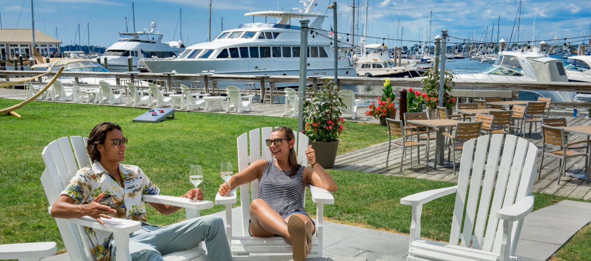 Two people relax on lawn chairs with a marina backdrop, enjoying drinks under a sunny sky near yachts and outdoor seating.