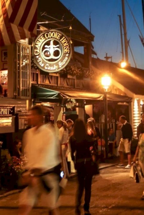 A lively evening street scene with people outside a café and restaurant, boats docked nearby, and glowing lights enhancing the atmosphere.