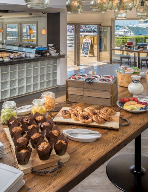 A buffet setup with pastries, muffins, fruit, yogurt, and plates on a wooden table in a modern dining area.