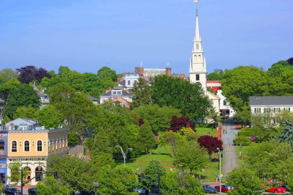 A quaint townscape with a park, charming buildings, a prominent church steeple, and lush greenery under a clear blue sky.