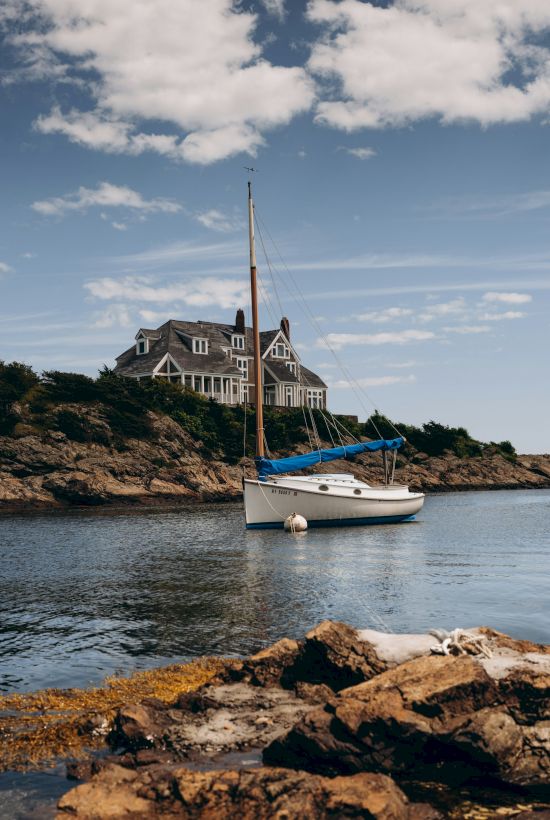 A sailboat on calm water, with a large house on a rocky shoreline under a partly cloudy sky.