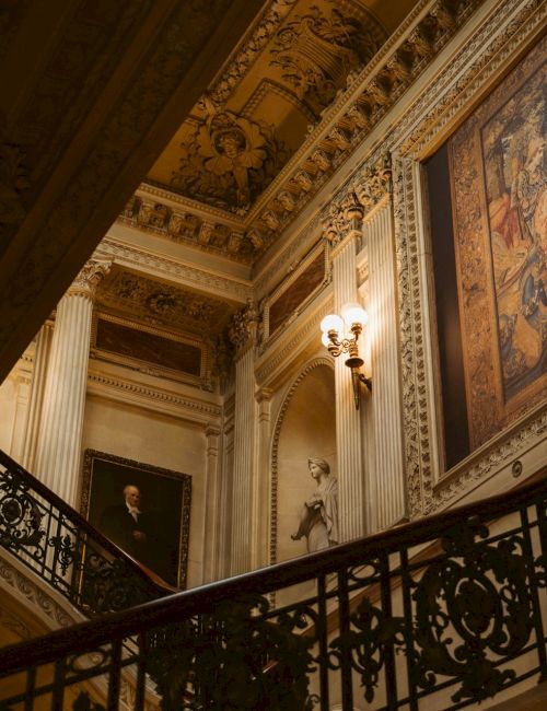 An ornate staircase with intricate railings, elaborate wall art, and vintage lighting in a grand, classical interior setting.