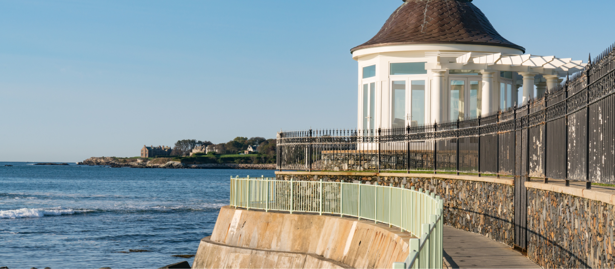 A seaside walkway with a railing leads to a gazebo by the ocean on a clear day.