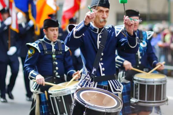 A marching band in traditional uniforms playing drums during a parade, with flags waving in the background.