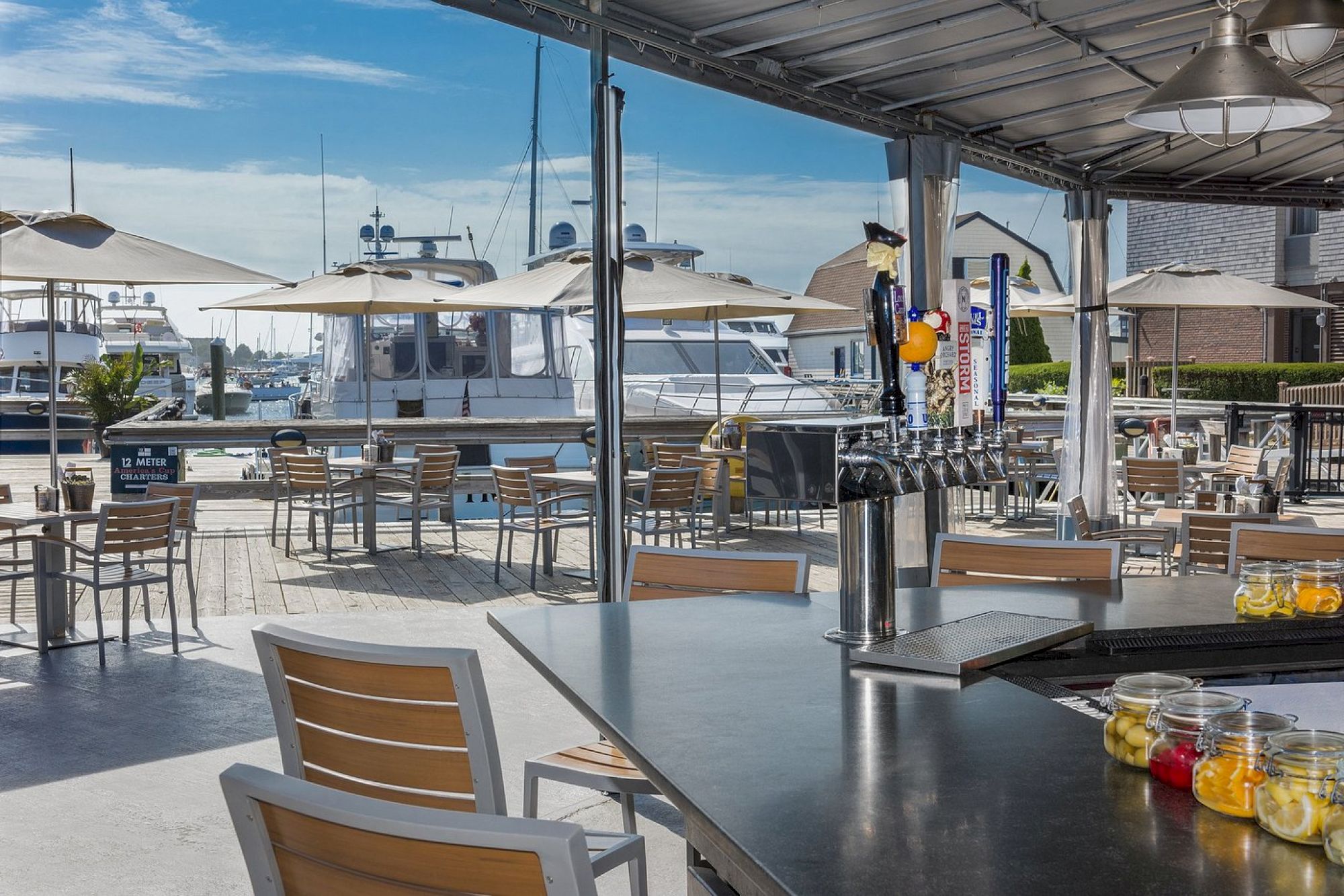 An outdoor bar and dining area by a marina, featuring wooden chairs, tables, draft beer taps, and boats in the background under sunny skies.