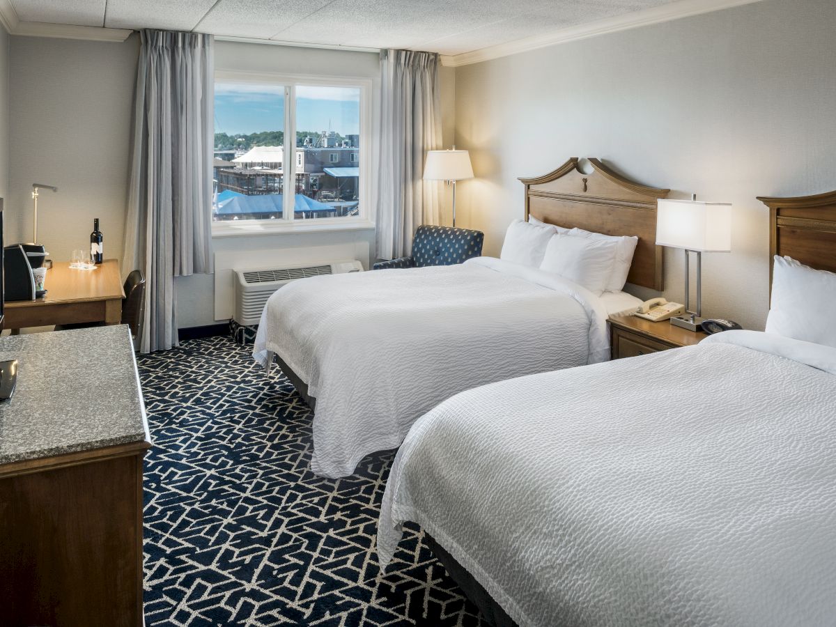 The image shows a hotel room with two beds, a TV on a wooden dresser, a desk with a lamp, and a window with curtains.